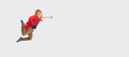 Portrait of little blond boy, pupil flying like super hero in red cape isolated on white studio background. Copyspace for ad. Childhood, education, emotion concept.の写真素材