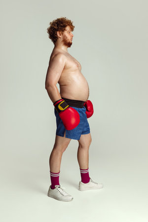 Funny red-headed man in blue boxing shorts and gloves isolated on gray studio background. Concept of sport, humor and body positive.の写真素材