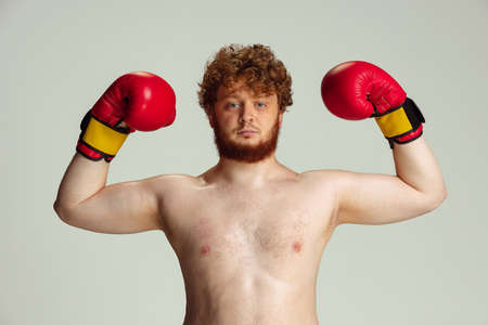 Funny red-headed man in blue boxing shorts and gloves isolated on gray studio background. Concept of...