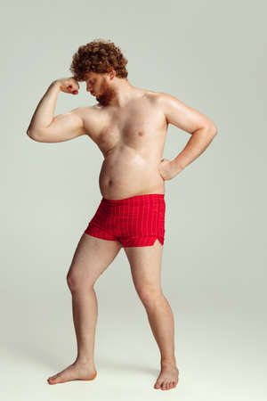 Cute red-headed man in red swimming shorts posing isolated on gray studio background. Concept of sport, humor and body positive.の写真素材