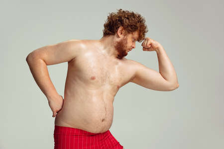 Cute red-headed man in red swimming shorts posing isolated on gray studio background. Concept of sport, humor and body positive.の写真素材