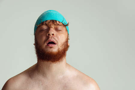 Close-up image of red-headed man in swimming hat posing isolated on gray studio background. Concept of sport, humor and body positive.の写真素材