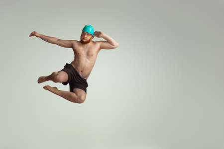 Funny dive. Cute red-headed man in red swimming shorts posing isolated on gray studio background. Concept of sport, humor and body positive.の写真素材