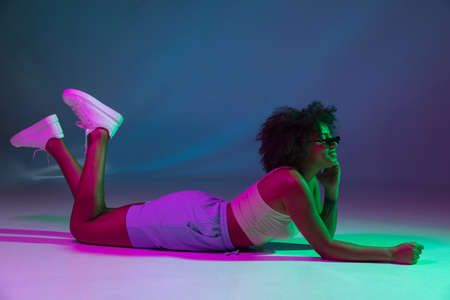 Portrait of African young girl posing isolated on dark blue studio background in pink neon light. Concept of human emotions, facial expression, youth, sales, ad.の写真素材