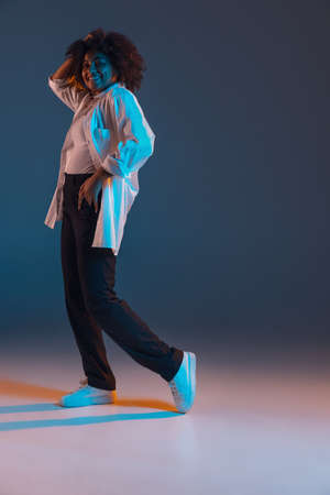 Full-length portrait of African young girl casual clothes, outfit isolated on dark blue studio background in pink neon light. Concept of human emotions, facial expression, youth, sales, ad.の写真素材