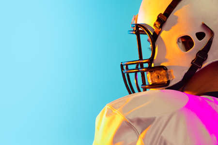 Cropped image of one American football player in sports equipment helmet and gloves isolated on blue studio background in neon light. Concept of sport, competitionの写真素材
