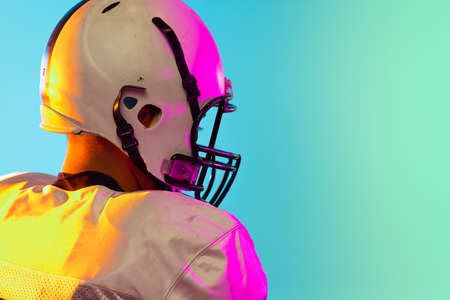 Cropped image of one American football player in sports equipment helmet and gloves isolated on blue studio background in neon light. Concept of sport, competitionの写真素材