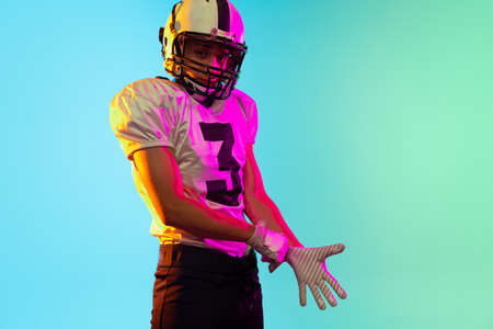 Portrait of one American football player in sports equipment helmet and gloves isolated on blue studio background in neon light. Concept of sport, competitionの写真素材