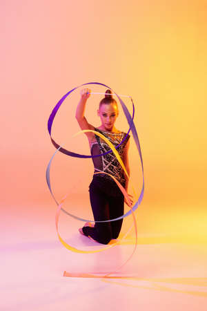 Portrait of little girl, rhythmic gymnastics artist isolated on pink yellow studio background in neon. Concept of sport, action, aspiration, education, active lifestyleの写真素材