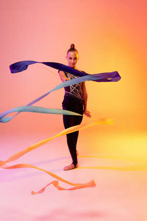 Portrait of little girl, rhythmic gymnastics artist isolated on pink yellow studio background in neon. Concept of sport, action, aspiration, education, active lifestyleの写真素材