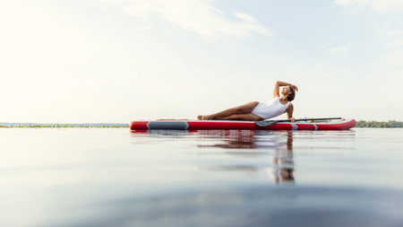 One young Caucasian beautiful slim girl sunbathe on summer evening on river on paddle board, SUP alone. Active life, sport, leisure activity conceptの写真素材