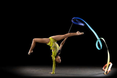 Slim sportive girl, rhythmic gymnastics artist in bright stage costume isolated on dark studio background in ray of light. Concept of sport, action, aspiration, active lifestyleの写真素材