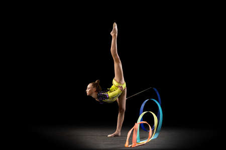 Slim sportive girl, rhythmic gymnastics artist in bright stage costume isolated on dark studio background in ray of light. Concept of sport, action, aspiration, active lifestyleの写真素材