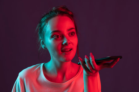 Portrait of pretty romantic girl with phone, gadgets isolated on dark purple studio background in red neon light, filter. Concept of human emotions, facial expression.の写真素材