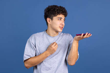Portrait of young curly man using phone isolated over blue studio background with copyspace for ad. Concept of human emotions, fashion, beauty, youthの写真素材