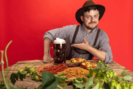 Happy smiling man dressed in traditional Austrian or Bavarian costume sitting at table with festive food and beer isolated over red backgroundの写真素材