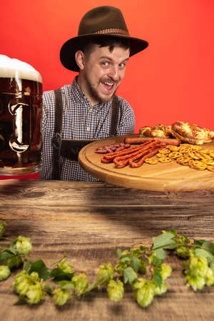 Happy smiling man dressed in traditional Austrian or Bavarian costume sitting at table with festive food and beer isolated over red backgroundの写真素材