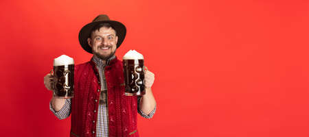 Happy smiling man dressed in traditional Austrian or Bavarian costume holding two mugs, glasses of dark black beer isolated over red background. Flyerの写真素材