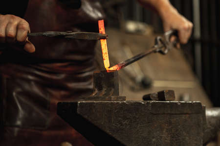 Close-up working powerful hands of male blacksmith forge an iron product in a blacksmith. Hammer, red hot metal and anvil. Concept of labor, retro professionsの写真素材