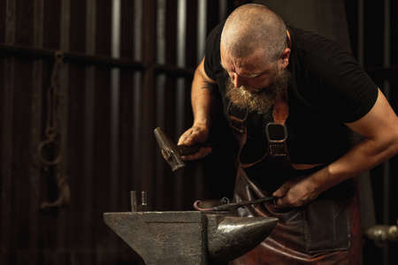 Bearded man, blacksmith working on creating handmade metal product at family smithy. Concept of labor, retro professions, power, beautyの写真素材