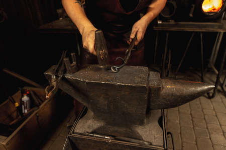 Close-up working powerful hands of male blacksmith forge an iron product in a blacksmith. Hammer,...