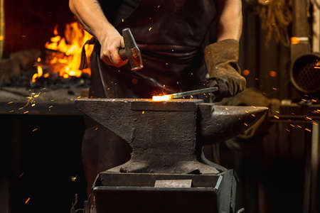 Bearded man, blacksmith manually forging the molten metal on the anvil in smithy with spark fireworks. Concept of labor, retro professions, family businessの写真素材