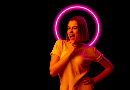 Portrait of young joyful girl isolated on dark studio background in multicolored neon light with geometric luminescent shape cirlce. Neon glowの写真素材