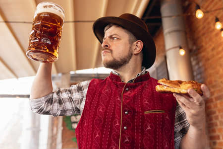 Portrait of happy bearded man in traditional Bavarian festive costume, hat and red vest tasting beer and snack during beer festival at bar, cafe, pub. Oktoberfest, october, autumnの写真素材