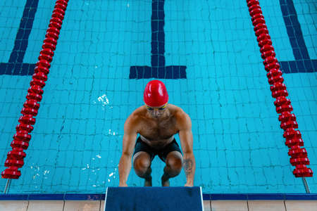 At start. Sportive male swimmer in red cap and goggles in motion and action during training at pool, indoors. Healthy lifestyle, power, energy, sports movement conceptの写真素材