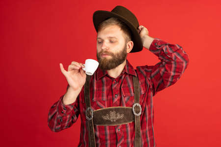 Comic portrait of bearded man, waiter in hat and traditional Bavarian costume isolated over red studio background. Oktoberfest, fest, holidays conceptの写真素材