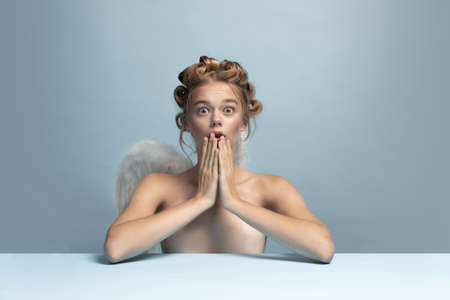 Portrait of young beautiful girl in image of angel with wings sitting and looking at camera closing mouth with hands isolated on blue gray studio background.の写真素材