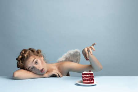 Portrait of young beautiful girl in image of angel with wings sitting isolated on blue gray studio background. Concept of beauty, purity, tenderness, inner worldの写真素材