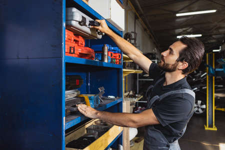 At car service station. One young man, male auto mechanic in dungarees working at car service...