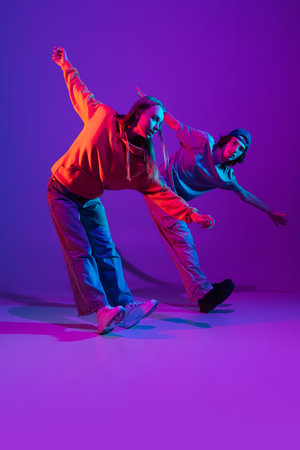Two dancers, young man and woman dancing hip-hop in casual sports youth clothes on gradient purple pink background at dance hall in neon light.の写真素材