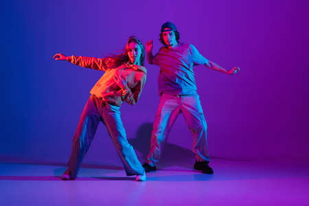Young man and woman dancing hip-hop in casual sports youth clothes on gradient purple pink background at dance hall in neon light.の写真素材