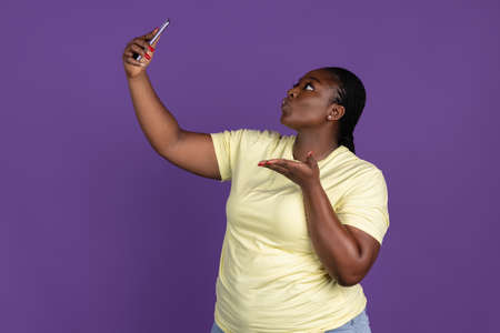 One African pretty woman with phone isolated on purple, lilac color studio background. Concept of human emotions, facial expression, natural beauty, bodypositiveの写真素材