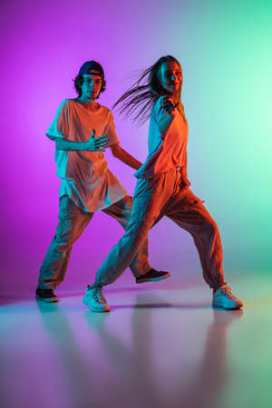 Young hip-hop dancers, stylish emotive girl and boy in action and motion in casual sports youth clothes on gradient multi colored background at dance hall in neon light.の写真素材