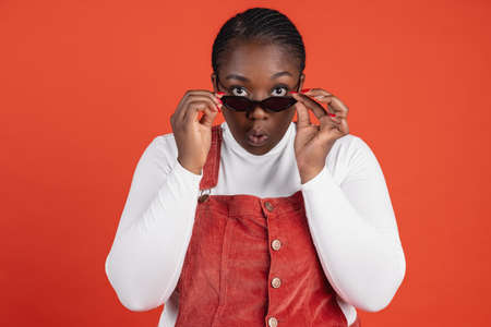 Bright portrait of African surprised woman in sunglasses isolated on red studio background. Concept of human emotions, facial expression, natural beauty, bodypositiveの写真素材