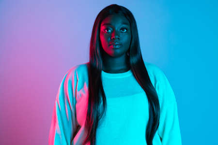 Close-up shot of African charming woman with long straight hair isolated on blue studio background in neon light. Concept of human emotionsの写真素材