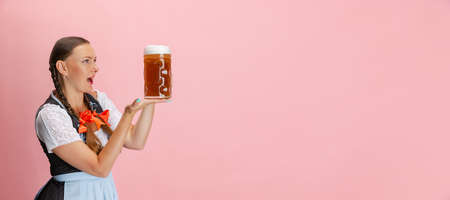 Flyer with adorable Oktoberfest woman, waitress wearing a traditional Bavarian or german dirndl holding one liter beer glass isolated on pink studio background.の写真素材