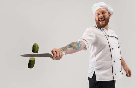 Unique cutting of vegetables. One hadsome bearded man, cook, male chef in white uniform cut cucumber isolated on white background.の写真素材