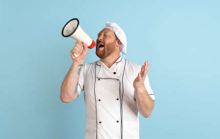 Comic portrait of hadsome bearded man, cook, male chef in white uniform shouting at megaphone isolated on blue background. Concept of job, occupation, humor.の写真素材