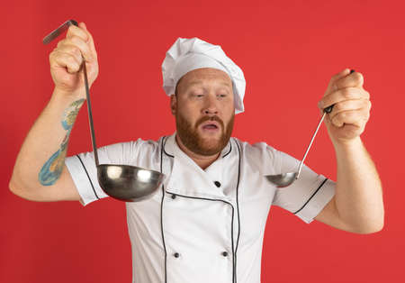 Comic portrait of hadsome bearded man, cook, male chef in white uniform having fun isolated on red...