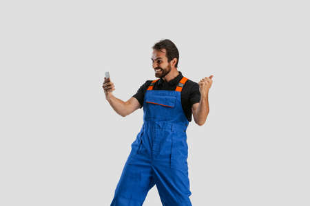 One young excited man, male auto mechanic or fitter in dungarees using phone isolated over white studio background. Concept of occupation, career, funny meme emotionsの写真素材