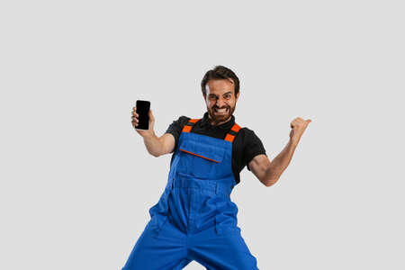 One young excited man, male auto mechanic or fitter in dungarees using phone isolated over white studio background. Concept of occupation, career, funny meme emotionsの写真素材
