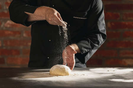Cropped image of male chef, cook in black kitchen uniform make bread dough at cafe, restaurant...