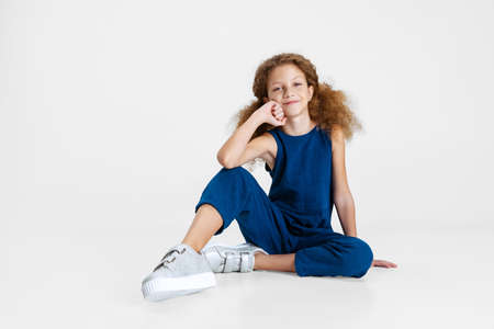 Portrait of little Caucasian cute girl in blue stylish jumpsuit sitting on floor isolated over white studio background.の写真素材