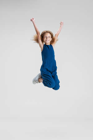 One little Caucasian cute girl in blue stylish jumpsuit jumping high and having fun isolated over white studio background.の写真素材