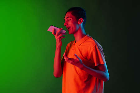 Portrait of young handsome man, student in casual clothes using phone isolated on green studio backgroud in neon light. Human emotions concept. Flyerの写真素材
