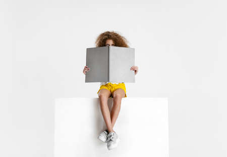 Portrait of little cute curly girl in casual clothes sitting on big box and reading book isolated on white studio background.の写真素材